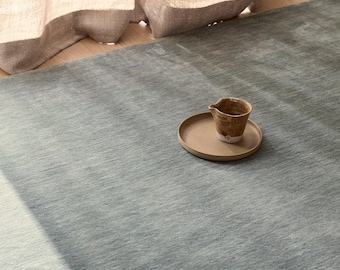 May include: A large, solid sage green area rug with a subtle, textured surface. Resting on the rug is a small, round, tan ceramic plate holding a single, small, brown, speckled ceramic pitcher with a spout and a slightly uneven, rustic finish.