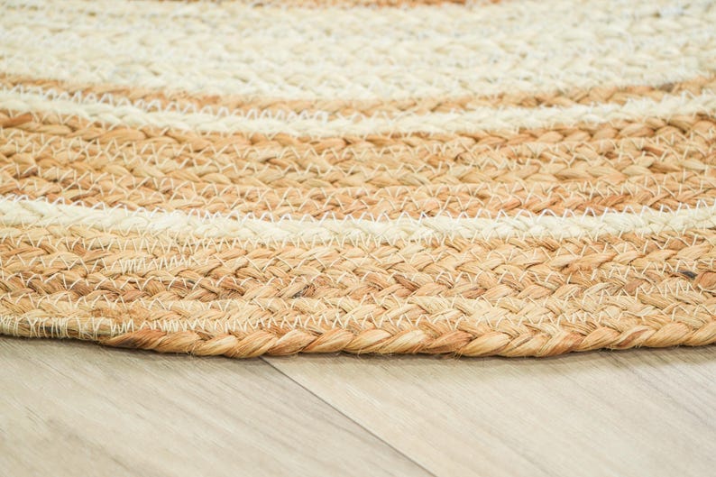 May include: A round braided rug featuring concentric circles of natural tan jute and off-white fibers. The texture is created by thick, tightly woven strands that form a repeating pattern of alternating light and neutral earth-toned bands.