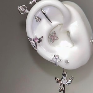 Könnte beinhalten: Eine Auswahl an silberfarbenen Ohrpiercings, darunter Schwerter, Schlüssel, Pfeil und Bogen sowie Engelsflügel-Designs, werden auf einem weißen Ohrmodell präsentiert. Einige Piercings weisen rosa Edelsteine auf. Der Schmuck ist vor einem neutralen Hintergrund platziert.