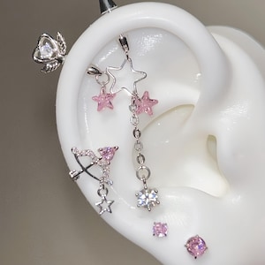 Könnte beinhalten: Ein silbernes Industrial-Barbell-Piercing mit einem geflügelten Herz, einem hängenden Stern-Anhänger und rosa Kristallen. Darunter ein silberner Pfeil-und-Bogen-Anhänger mit rosa Kristallen und einem kleinen Stern sowie zwei einzelne Ohrstecker.