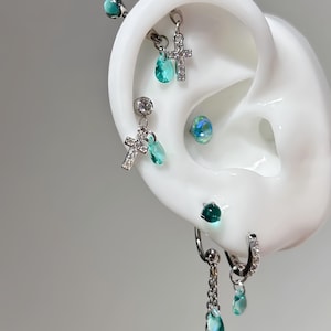 Puede incluir: Un surtido de pendientes y piercings plateados, que incluyen colgantes de cruz, gemas en forma de lágrima y una barra con púas. La joyería se exhibe en un modelo de oreja blanca, con un fondo gris.
