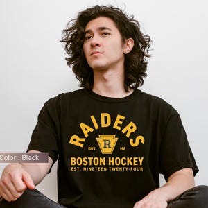 May include: Black t-shirt with the text "RAIDERS BOSTON HOCKEY EST. NINETEEN TWENTY-FOUR" in yellow. The shirt has a crew neck and short sleeves. The image shows a person wearing the shirt.