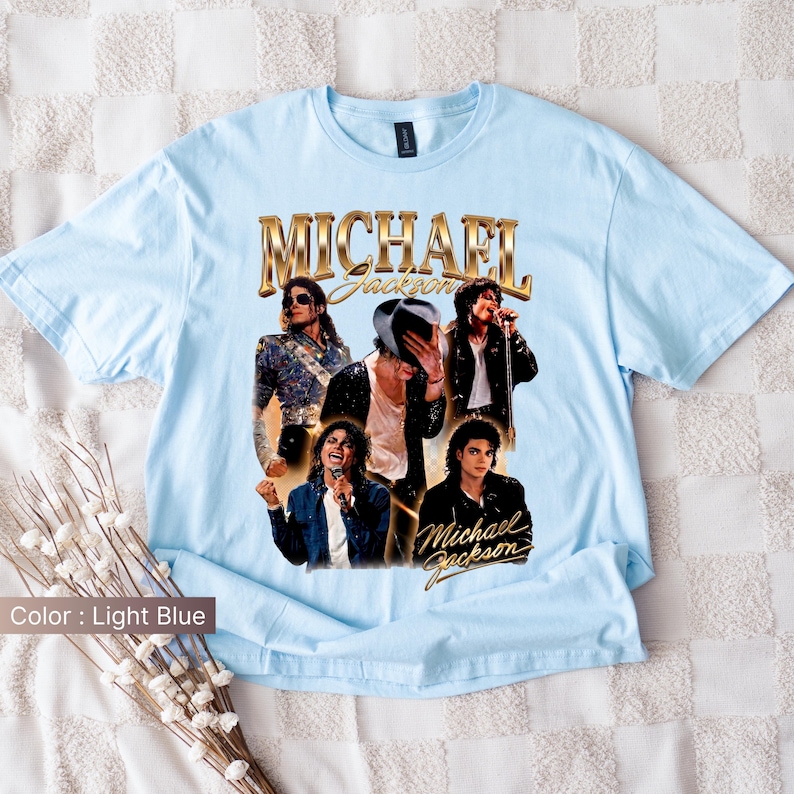 May include: Light blue t-shirt featuring a collage of images of Michael Jackson, with the name "Michael Jackson" in gold lettering. The shirt showcases various poses of the artist, capturing his iconic style and performances. The shirt is made of a soft material.