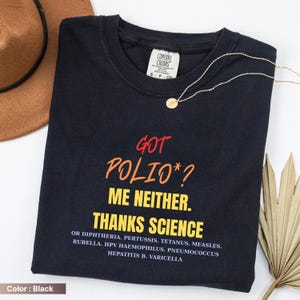 May include: Black t-shirt with the text "GOT POLIO*? ME NEITHER. THANKS SCIENCE" in red, orange, and yellow. The shirt also lists several diseases. A gold necklace and a brown hat are also in the image.