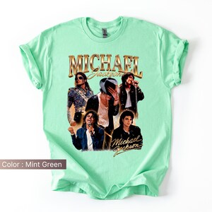 May include: Mint green t-shirt featuring a collage of images of Michael Jackson. The shirt has the words "MICHAEL Jackson" in gold lettering. The shirt is a crew neck and has rolled-up sleeves.