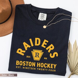 May include: Black t-shirt with the word "RAIDERS" in yellow arched text. Below, the text "BOSTON HOCKEY EST. NINETEEN TWENTY-FOUR" is printed in yellow. A gold necklace and a brown hat are also visible.