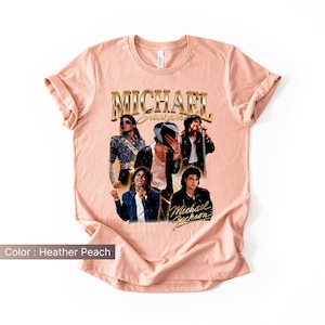 May include: Heather peach-colored t-shirt featuring a collage of Michael Jackson images. The shirt has the text "Michael Jackson" in gold lettering. The images show Jackson in various poses, wearing different outfits.