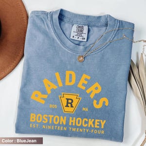 May include: Blue jean-colored t-shirt with the word "RAIDERS" in yellow block letters. Below the word "RAIDERS" is the text "BOSTON HOCKEY EST. NINETEEN TWENTY-FOUR". A gold necklace with a pendant is draped across the shirt.