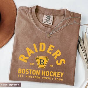 May include: Espresso-colored t-shirt with the text "RAIDERS BOSTON HOCKEY EST. NINETEEN TWENTY-FOUR" in yellow. The shirt has a crew neck and short sleeves. A silver necklace with a pendant is draped across the shirt.