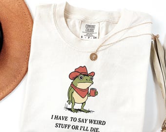 May include: Ivory-colored t-shirt featuring a cartoon frog wearing a cowboy hat and holding a cup. The text "I HAVE TO SAY WEIRD STUFF OR I'LL DIE" is printed below the frog. A gold necklace and a brown hat are also visible.