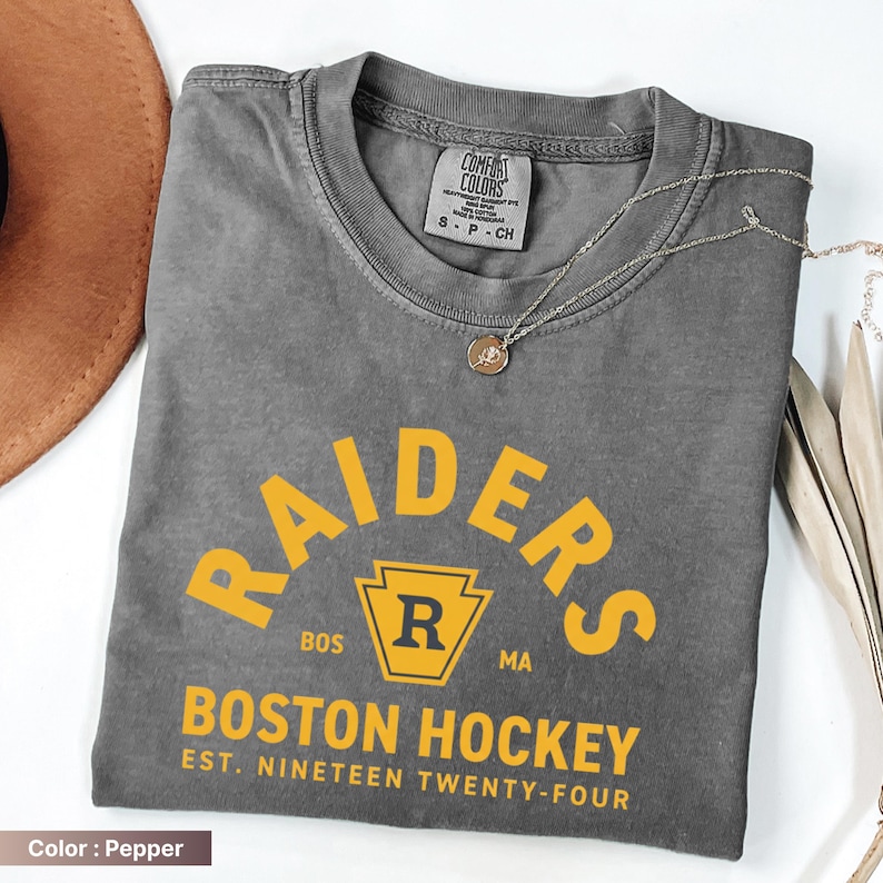 May include: A gray t-shirt with the word "RAIDERS" in yellow arched text. Below, the text "BOSTON HOCKEY EST. NINETEEN TWENTY-FOUR" is printed in yellow. A silver necklace with a pendant is draped on the shirt.