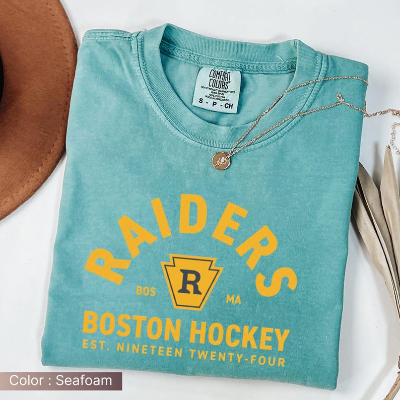 May include: Seafoam green t-shirt with "RAIDERS" arched text in yellow, and "BOSTON HOCKEY" below. The shirt includes a yellow and black emblem with the letter "R". The text "EST. NINETEEN TWENTY-FOUR" is also visible. A gold necklace is draped across the shirt.
