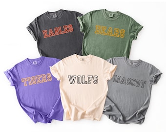 May include: Five short-sleeved t-shirts in various colors: dark gray, green, purple, cream, and light gray. Each shirt features a different word in a collegiate-style font: "EAGLES", "BEARS", "TIGERS", "WOLFS", and "MASCOT".