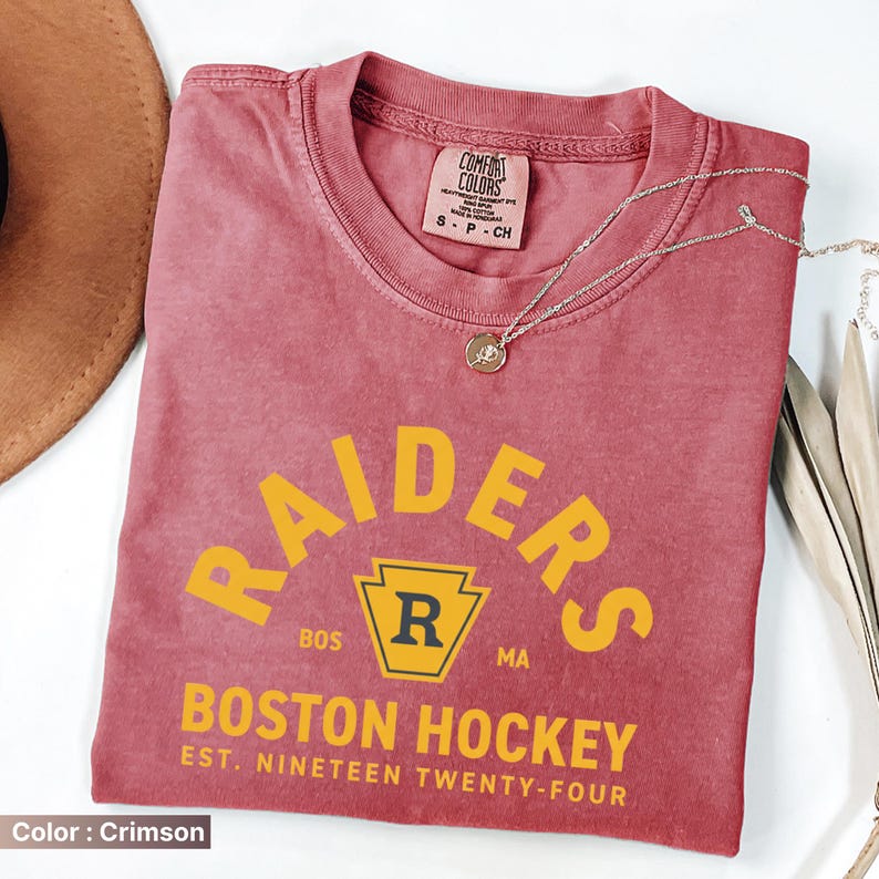 May include: Crimson-colored t-shirt with the word "RAIDERS" in yellow arched across the top. Below, the text "BOSTON HOCKEY EST. NINETEEN TWENTY-FOUR" is printed in yellow. A silver necklace with a pendant is draped across the shirt.