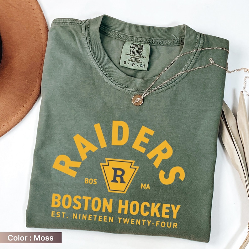 May include: Moss green t-shirt with "RAIDERS" arched text in yellow, and "BOSTON HOCKEY" below. The shirt includes a gold pendant necklace. The shirt is labeled "COMFORT COLORS" and "S-P-CH".