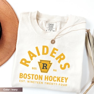 May include: Ivory-colored t-shirt with the text "RAIDERS BOSTON HOCKEY EST. NINETEEN TWENTY-FOUR" in yellow. The shirt has a crew neck and short sleeves. A gold necklace with a pendant is draped across the shirt.