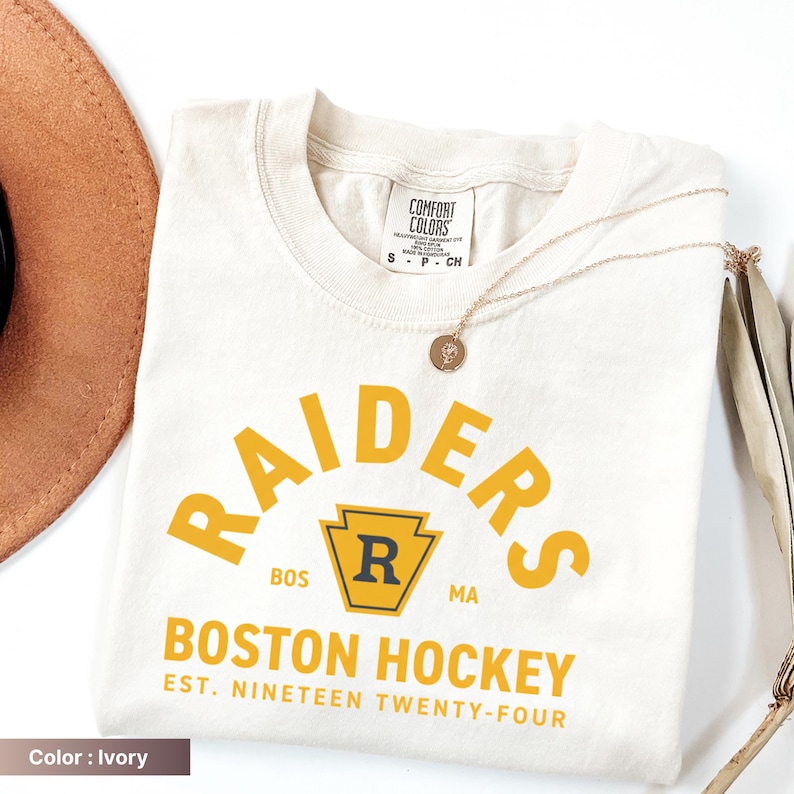 May include: Ivory-colored t-shirt with the text "RAIDERS BOSTON HOCKEY EST. NINETEEN TWENTY-FOUR" in yellow. The shirt has a crew neck and short sleeves. A gold necklace with a pendant is draped across the shirt.