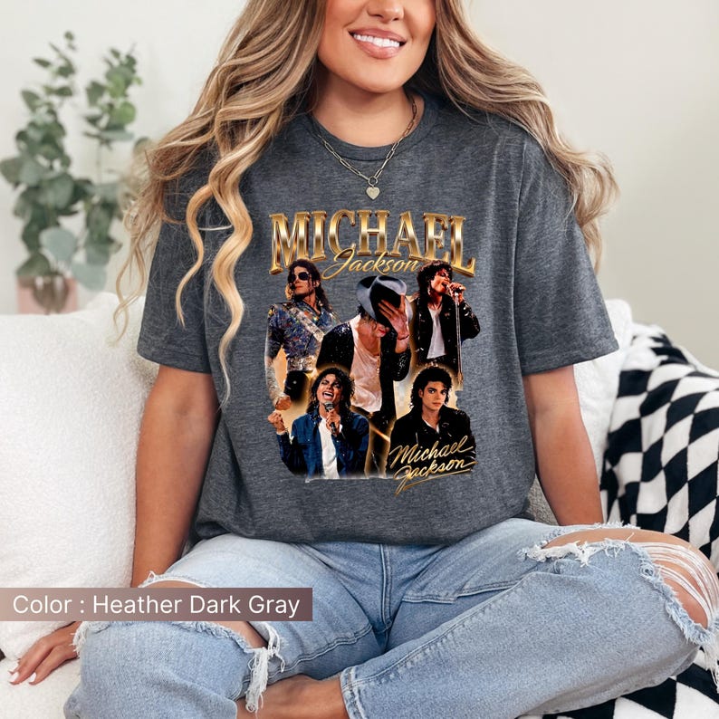 May include: Heather dark gray t-shirt featuring a collage of images of Michael Jackson. The shirt has the name "Michael Jackson" in gold lettering at the top. The shirt is a crew neck and is worn by a person.