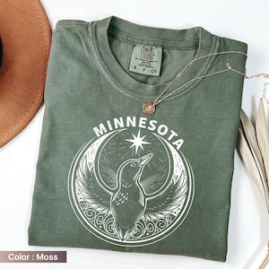 May include: A moss-colored t-shirt with the word "MINNESOTA" above a graphic of a bird within a crescent moon and star design. A gold necklace with a pendant rests on the shirt. The shirt has a crew neck and short sleeves.