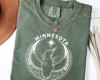 May include: A moss-colored t-shirt with the word "MINNESOTA" above a graphic of a bird within a crescent moon and star design. A gold necklace with a pendant rests on the shirt. The shirt has a crew neck and short sleeves.