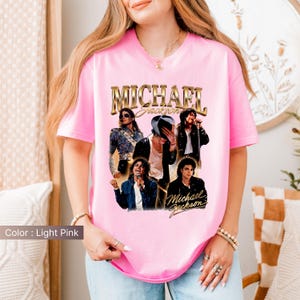 May include: Light pink t-shirt featuring a gold-colored "Michael Jackson" graphic. The graphic includes multiple images of Michael Jackson. The shirt is a crew neck and has short sleeves.