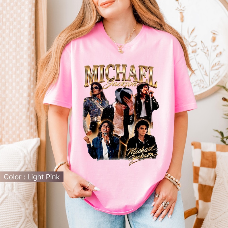 May include: Light pink t-shirt featuring a gold-colored "Michael Jackson" graphic. The graphic includes multiple images of Michael Jackson. The shirt is a crew neck and has short sleeves.