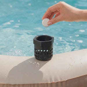 Peut inclure: Un panier de filtre de spa noir avec un dessus hexagonal et des côtés perforés. Un comprimé blanc est placé dans le panier. Le panier est au bord d'un spa avec de l'eau bleue en arrière-plan.