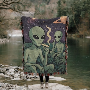 May include: A woven throw blanket featuring a space-themed design with two green aliens smoking. The background is a dark purple with stars, a galaxy, and a planet. The blanket has a fringed edge.