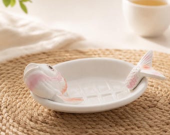 May include: A white ceramic dish shaped like a fish, featuring a sculpted fish head on one end and a tail on the other. The interior of the dish has a grid pattern embossed on the bottom, and the fish head and tail are accented with soft pink.