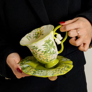 May include: A ceramic teacup and saucer set featuring a cream-coloured interior and a vibrant green exterior. The surface is decorated with detailed botanical illustrations of yellow flowers and green leaves. The handle is shaped like a lily of the valley.