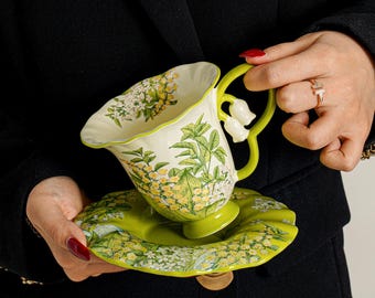 May include: A ceramic teacup and saucer set featuring a cream-coloured interior and a vibrant green exterior. The surface is decorated with detailed botanical illustrations of yellow flowers and green leaves. The handle is shaped like a lily of the valley.