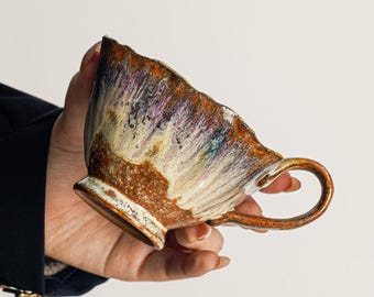 May include: A ceramic teacup featuring a textured, uneven rim and a curved handle. The exterior displays a reactive glaze with blended streaks of deep brown, metallic copper, creamy white, and hints of purple, creating a mottled, organic appearance.