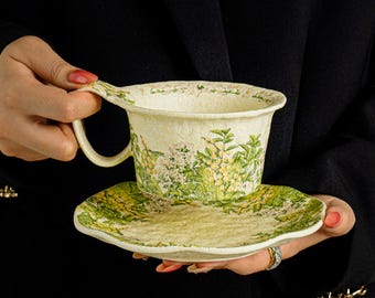 May include: A cream-coloured ceramic teacup and saucer set featuring a textured surface and a detailed botanical print of yellow and white flowers with green leaves. The teacup has a wide, flared rim and a curved handle, while the saucer has a scalloped edge.