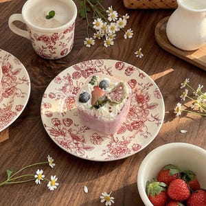 May include: A still-life image featuring a heart-shaped dessert on a floral plate, a cup of coffee, and a bowl of strawberries. The plate and cup have a red floral pattern. Daisies and a small pitcher are also present.