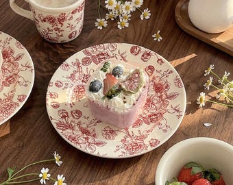 May include: A table setting with a heart-shaped dessert on a floral plate, a cup of coffee, and a bowl of strawberries. The plate and cup have a red floral pattern. Daisies and a small jug are also present.