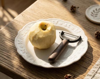 May include: A peeled apple on a white plate with a decorative edge, alongside a peeler with a dark wood handle. The plate has embossed floral patterns. The scene is set on a wooden table, suggesting a kitchen or dining setting.