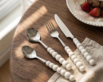 May include: A stainless steel cutlery set with white, beaded handles. The set includes a spoon, a smaller spoon, a fork, and a knife. The cutlery is arranged on a wooden table next to a plate of food.