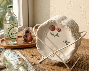 May include: A set of white ceramic plates with a floral design, held in a white metal rack. The plates have a scalloped edge and feature pink flowers and green leaves. A bottle of soda water and a small jar are also visible.