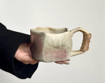 May include: A ceramic mug with an irregular, organic shape and a textured, mottled finish in shades of cream, grey, and muted pink. The mug features a square-like rim and a handle wrapped in natural-coloured twine, providing a tactile grip.