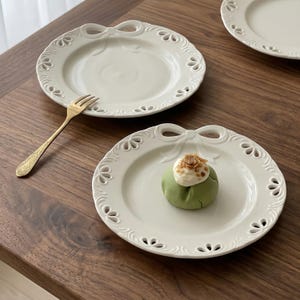 May include: White ceramic plates with a decorative bow design on the rim. One plate holds a green pastry topped with cream and sprinkles. A gold fork rests on the wooden surface, next to the plates.