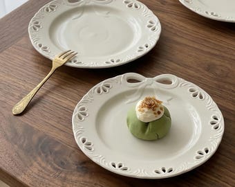 May include: White ceramic plates with a decorative bow design on the rim. One plate holds a green pastry topped with cream and sprinkles. A gold fork rests on the wooden surface, next to the plates.