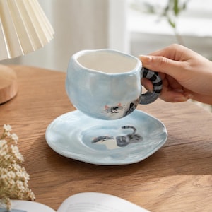 May include: A light blue ceramic cup and saucer set with an irregular, wavy rim. The cup features a painted cat face and a striped handle, while the saucer displays a matching cat illustration. Both pieces have a glossy, hand-crafted appearance.