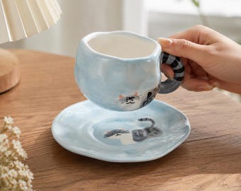 May include: A light blue ceramic cup and saucer set with an irregular, wavy rim. The cup features a painted cat face and a striped handle, while the saucer displays a matching cat illustration. Both pieces have a glossy, hand-crafted appearance.
