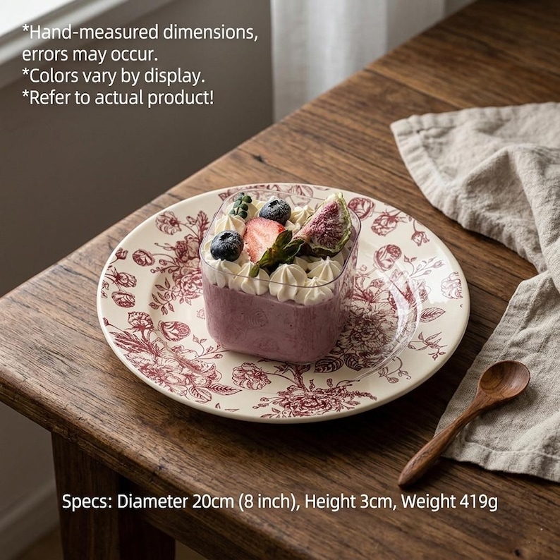 May include: A dessert with whipped cream, blueberries, and a strawberry slice sits atop a floral patterned plate. The plate is 8 inches in diameter. The dessert is in a clear square container. A wooden spoon and a linen napkin are nearby. The dessert is 3cm high and weighs 419g.