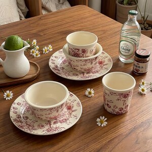 May include: A set of cream-colored ceramic tableware with a red floral pattern. The set includes bowls, saucers, and a cylindrical cup. A white pitcher with a lime inside is also present, along with a bottle of soda water and a jar of food.