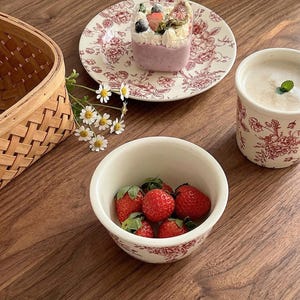 May include: A still-life image featuring a bowl of fresh strawberries, a dessert in a clear cup, and a floral-patterned plate and mug. The items are arranged on a wooden surface, with a woven basket and flowers adding to the aesthetic.