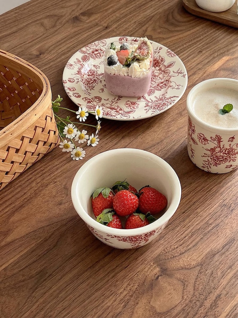 May include: A still-life image featuring a bowl of fresh strawberries, a dessert in a clear cup, and a floral-patterned plate and mug. The items are arranged on a wooden surface, with a woven basket and flowers adding to the aesthetic.