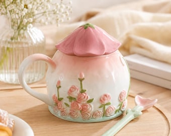May include: A ceramic mug with a lid shaped like a pink flower petal, featuring a white base decorated with embossed pink roses and green stems. The mug includes a matching spoon with a light green handle and a pink flower-shaped tip.