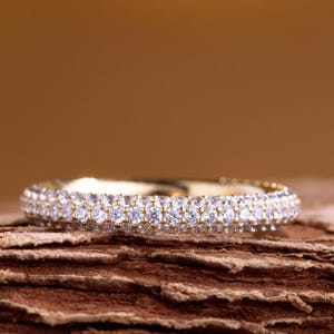 May include: A gold band ring with two rows of small, clear gemstones. The ring is set against a brown, textured background, creating a contrast between the shiny ring and the rustic backdrop. The ring is a piece of jewelry.