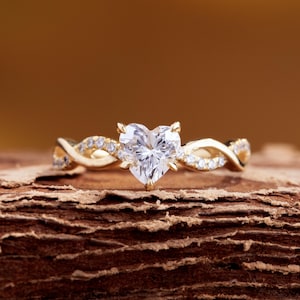 May include: A gold ring with a heart-shaped diamond center stone. The band features a twisted design with smaller accent diamonds. The ring is set on a textured, brown surface, with a warm, golden background.